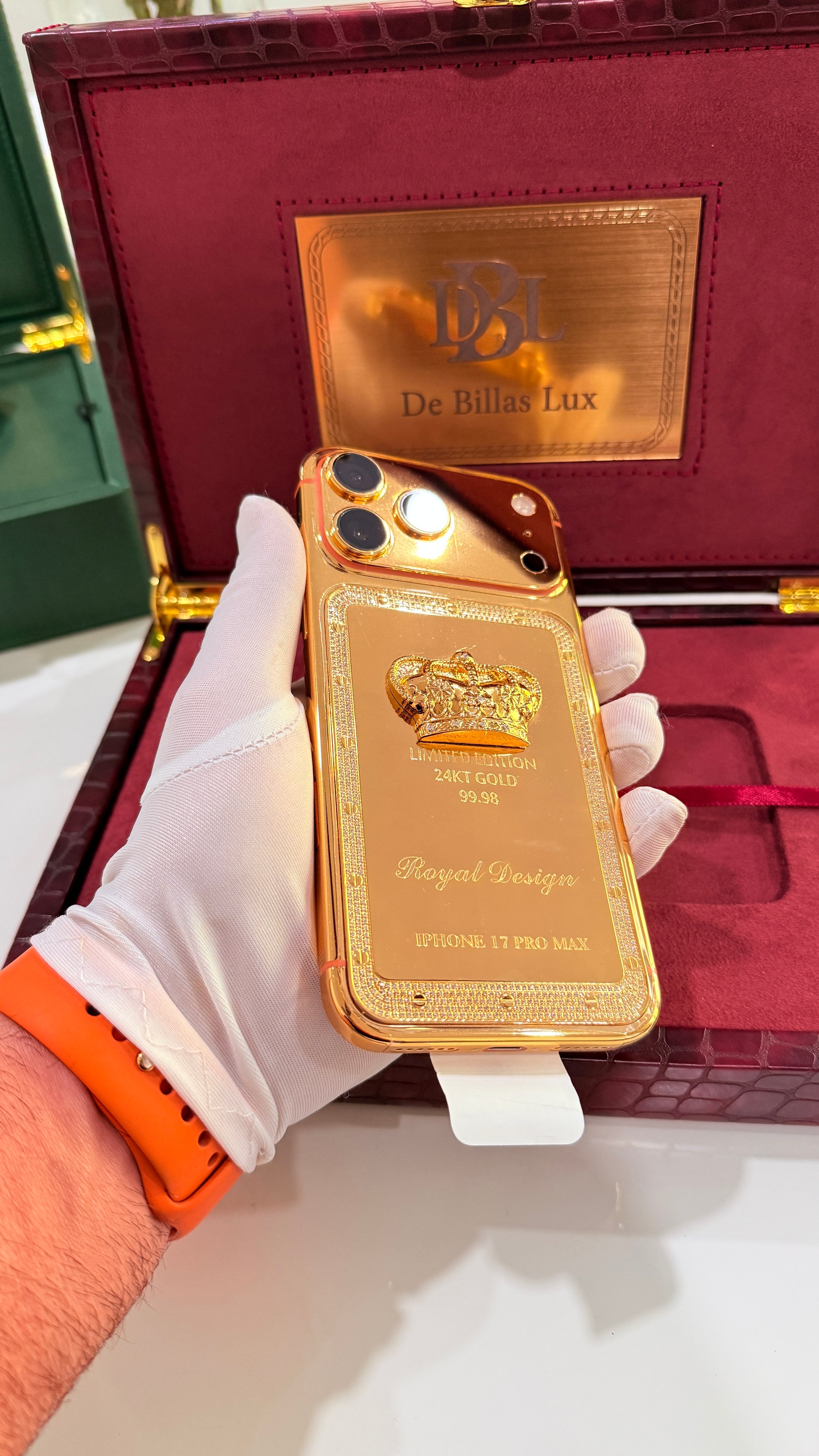 A gold-plated luxury phone case featuring a crown emblem and 'Royal Design' text, held by a hand wearing a white glove in front of its dark red and gold De Bellois Lux presentation box.